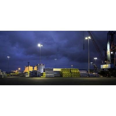 Industrial site at night with stacked metal bars and timber materials on an illuminated harbour. A crane is visible in the background.