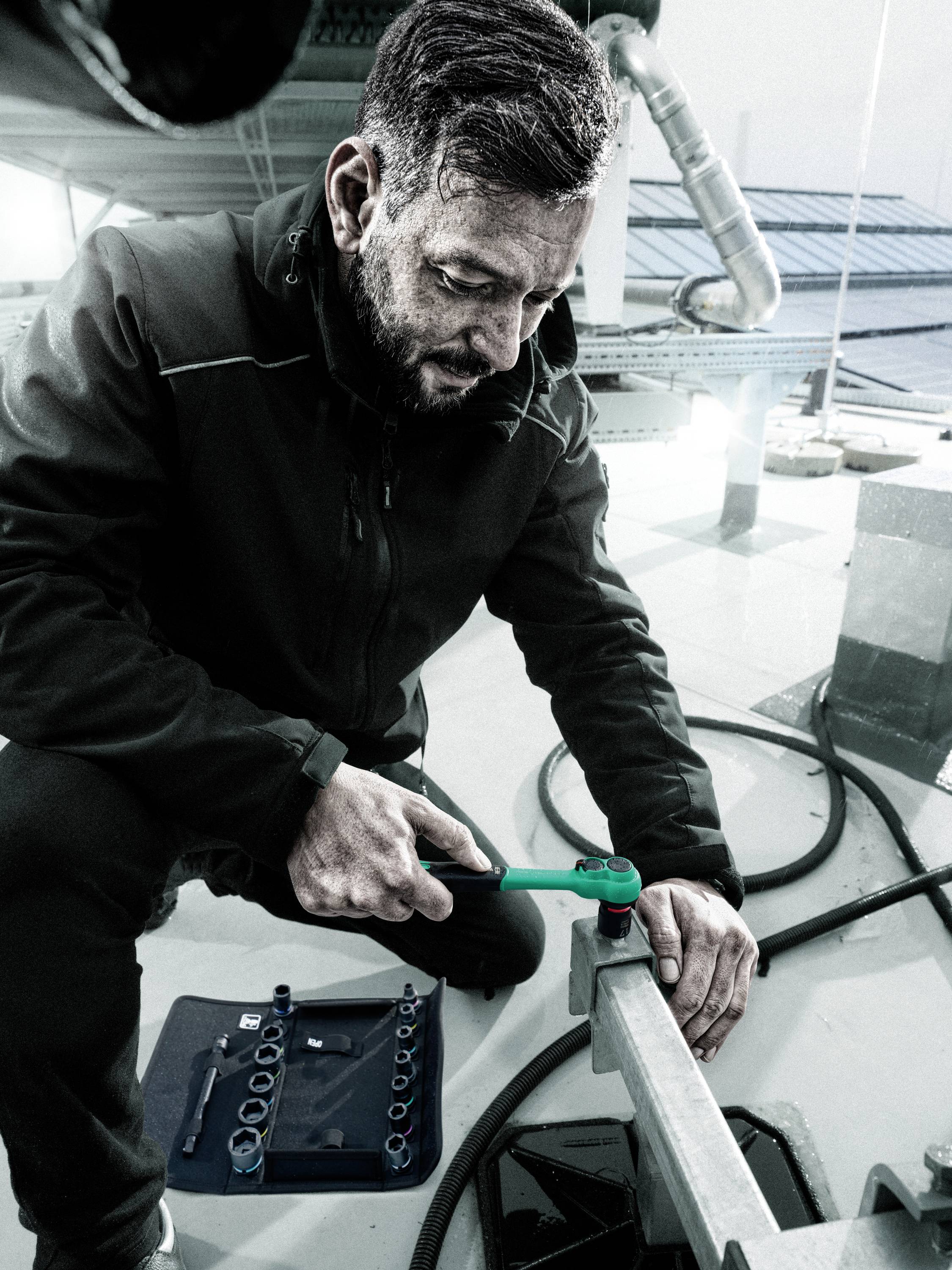 A man in workwear is tightening a screw with a green adjustable spanner. A toolbox lies beside him.