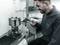 A man is measuring a machine part with a digital measuring instrument in a laboratory or workshop. A computer is standing in the background.