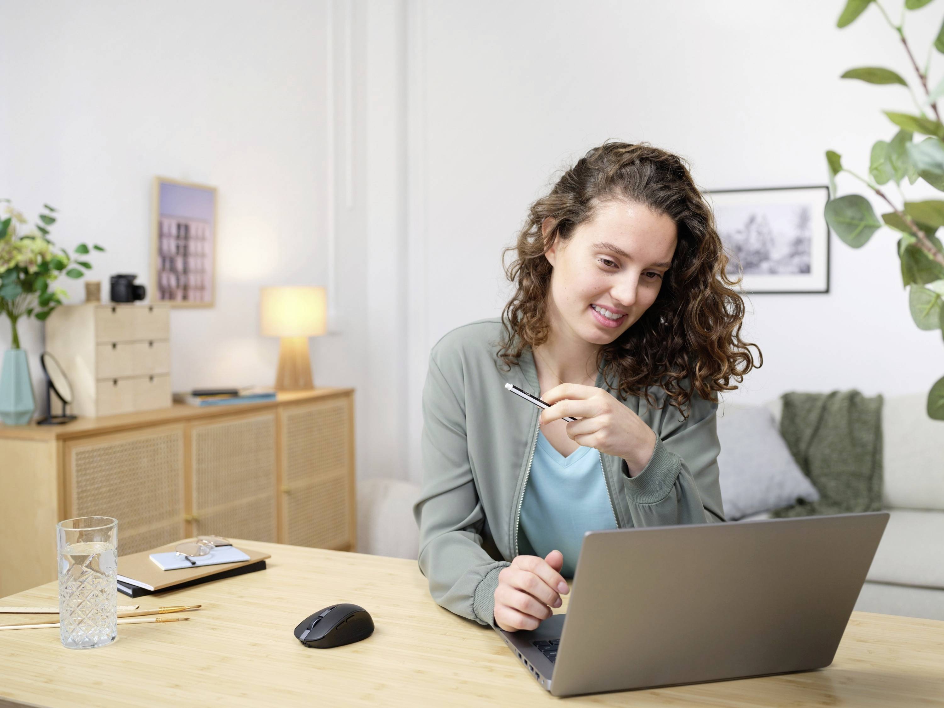 A woman with curly hair sits at a table using a laptop, smiling. Plants and pictures are visible in the background.