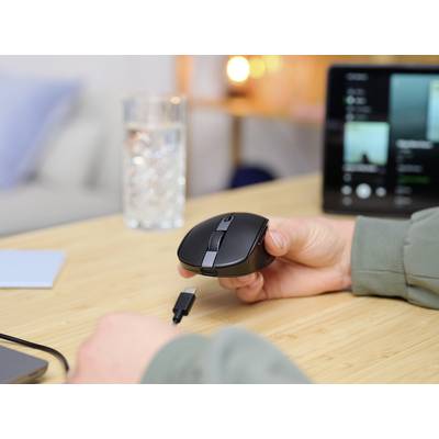 A person is holding a black computer mouse, ready to connect a USB cable. A glass of water and a screen are in the background.