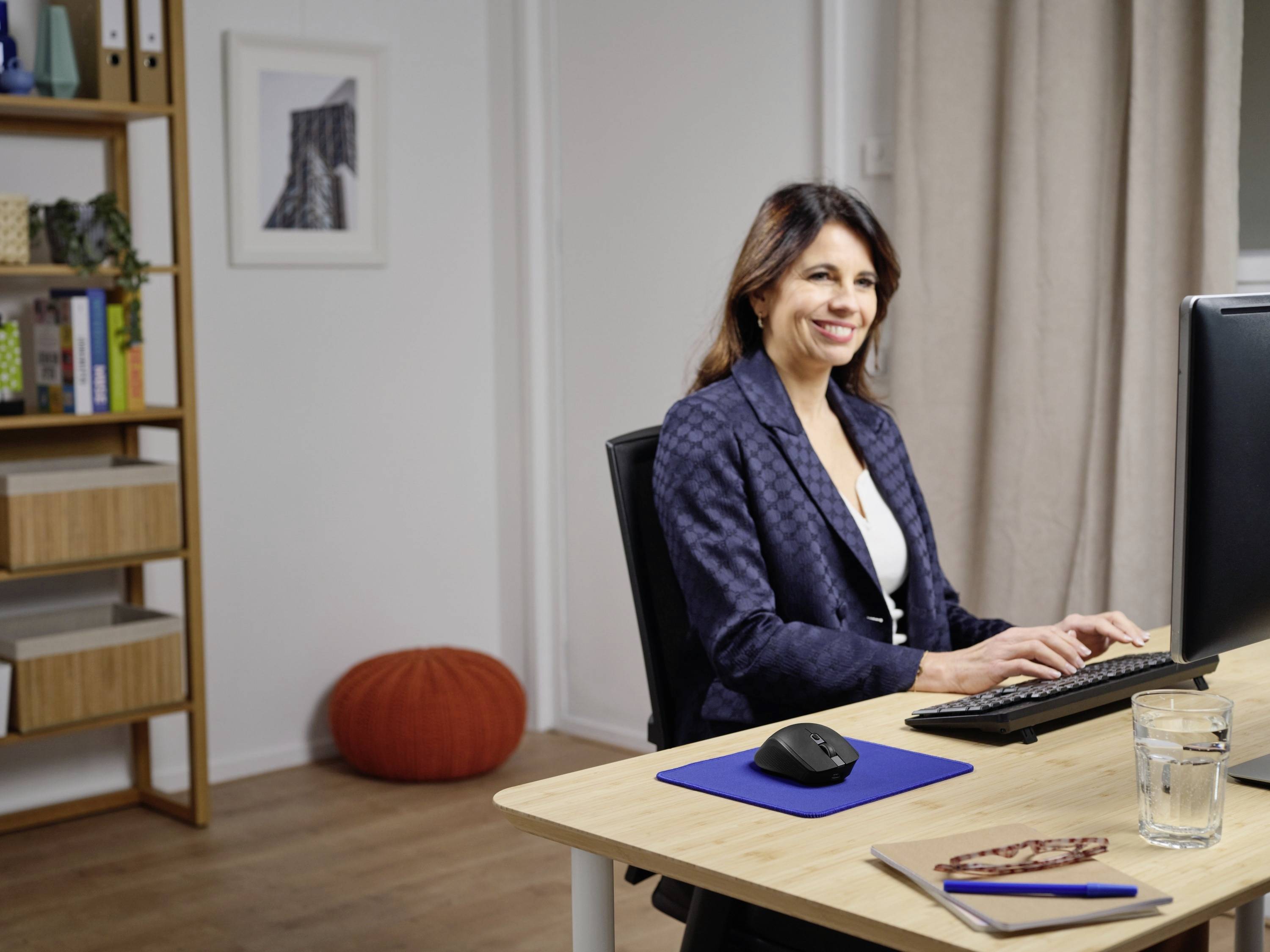 A woman is sitting at a desk and working on a computer. In the background, a bookshelf and an orange cushion are visible.