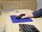 A hand operates a black computer mouse on a blue mouse mat, resting on a wooden table. A computer keyboard is visible in the background.