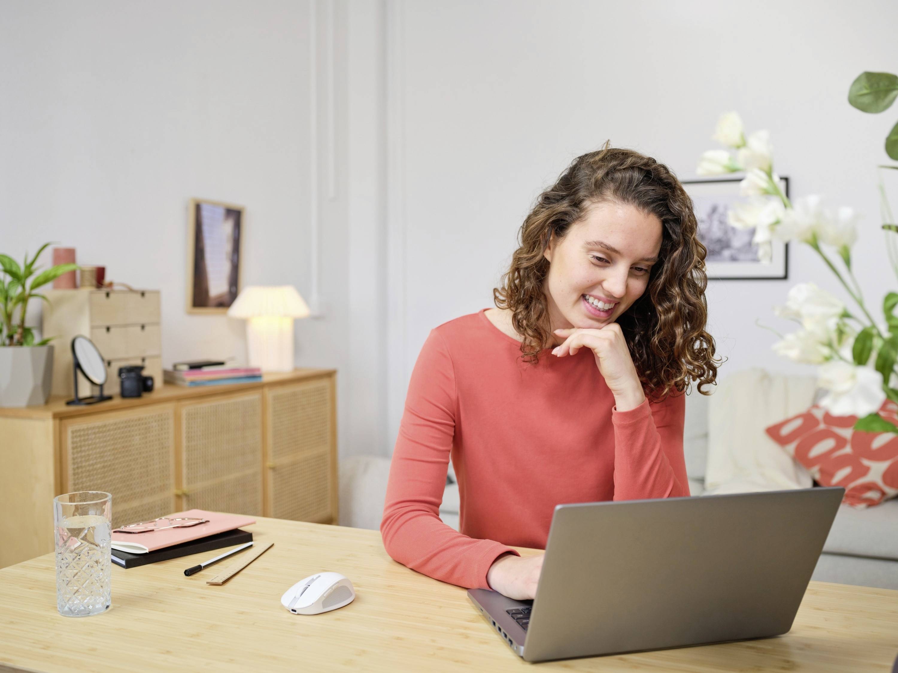 A smiling person is sitting at a table, working on a laptop. Plants and pictures can be seen in the background.