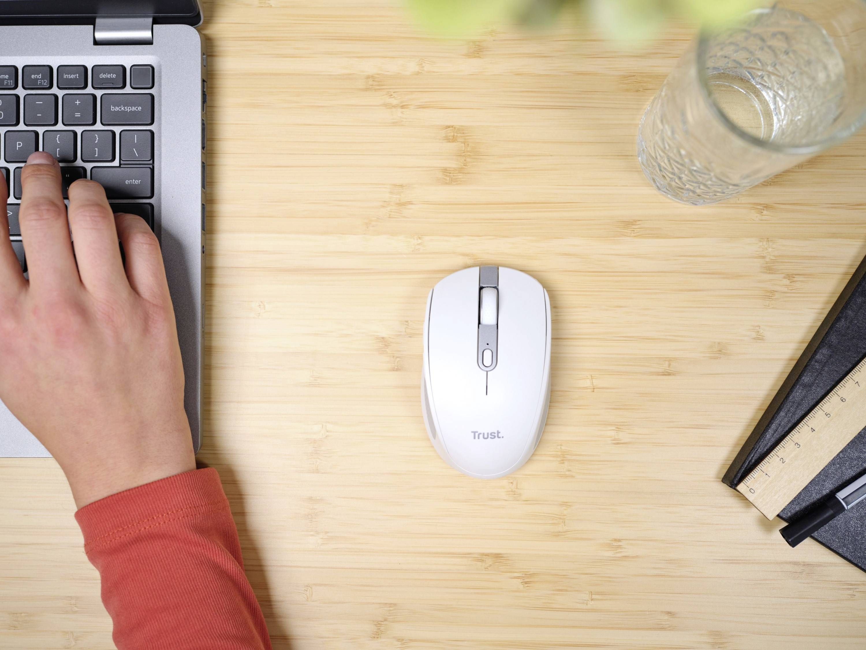 A hand is typing on a laptop keyboard. Beside it, a computer mouse, a glass of water and a ruler are lying on a wooden desk.