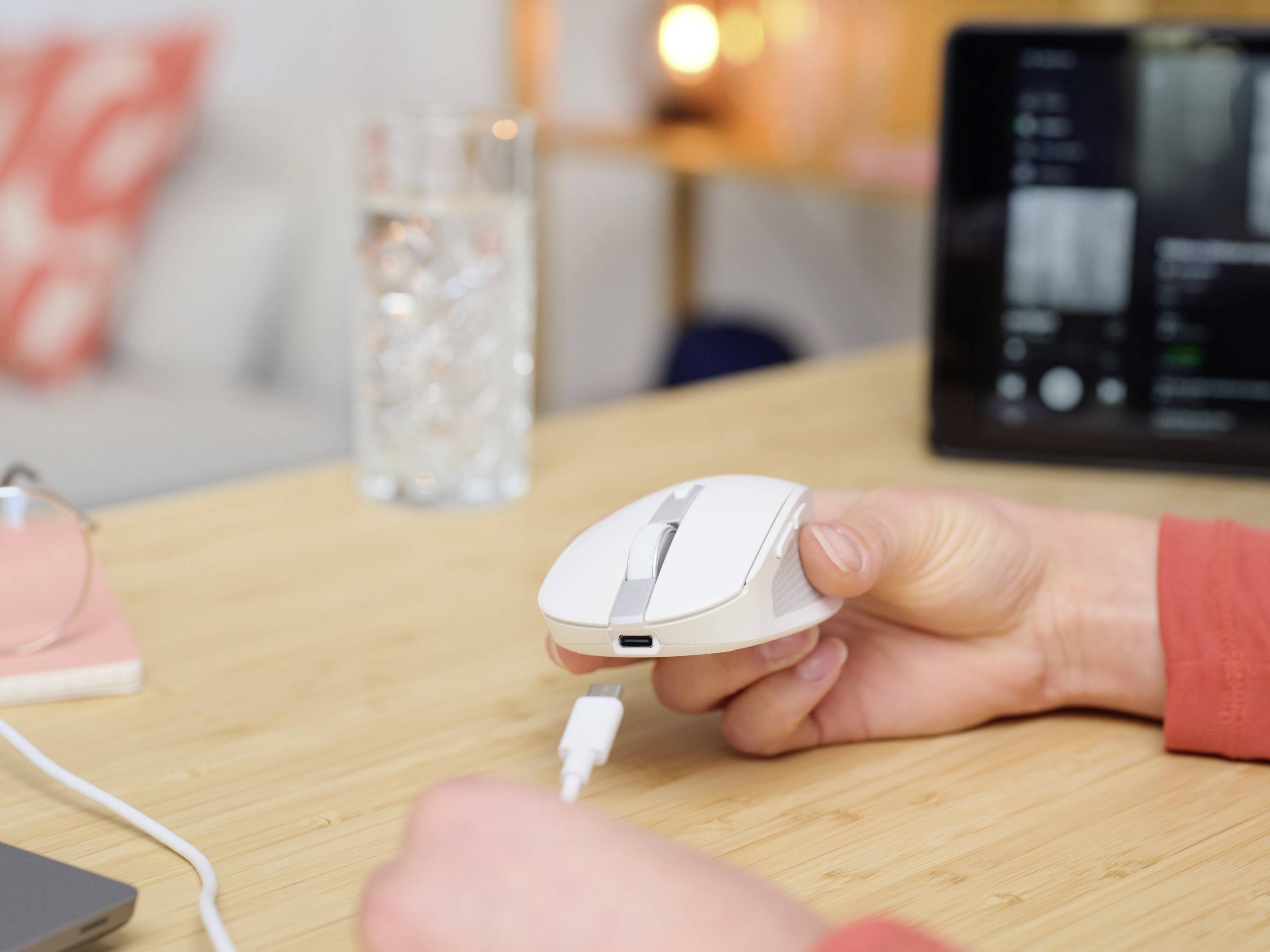 A hand is holding a computer mouse, which is connected via a USB cable on a desk. A glass of water sits in the background.