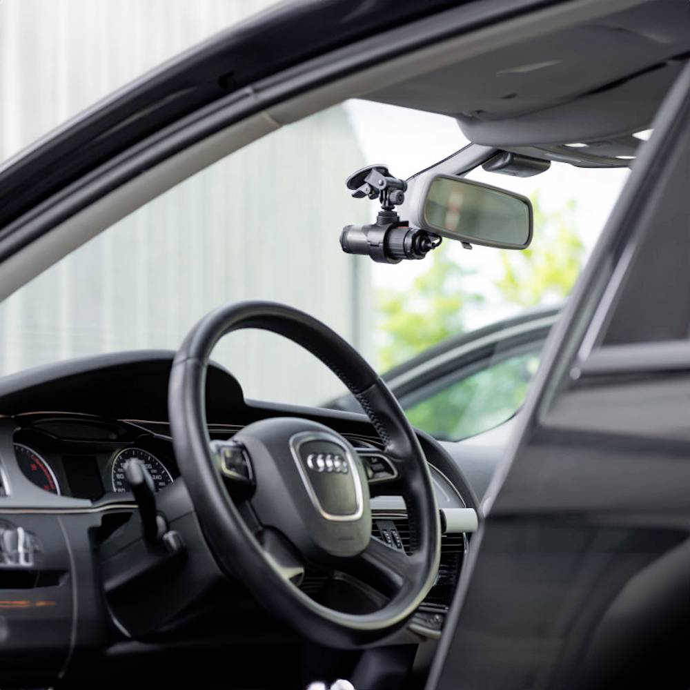 Interior view of a car with a dashcam mounted on the rear-view mirror, facing the road. Steering wheel and dashboard are visible.