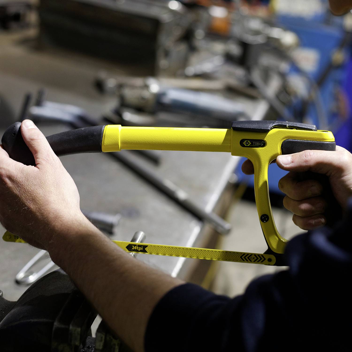 A person is sawing a piece of metal with a yellow metal saw in a workshop. Tools and equipment are visible in the background.