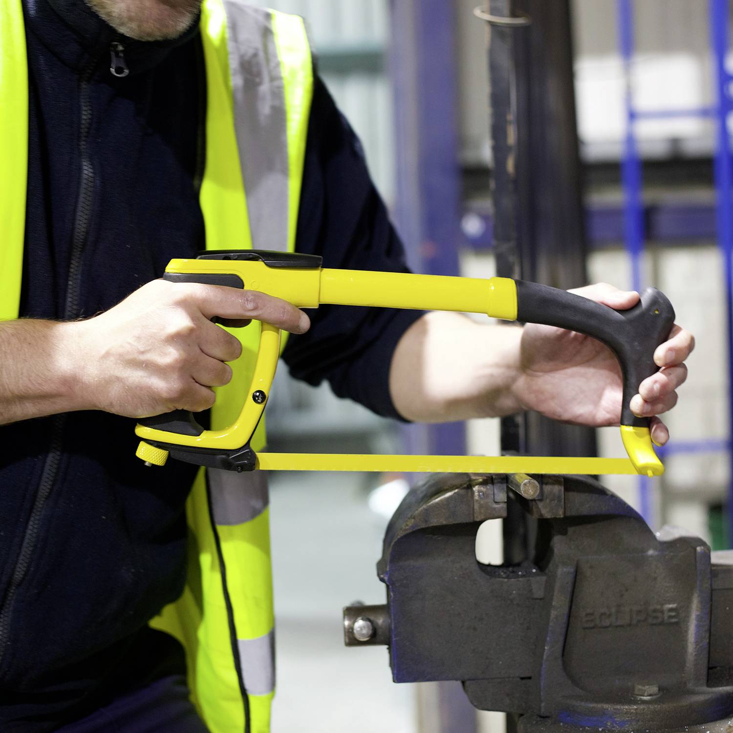 A person in a yellow hi-vis vest is using a hacksaw to cut metal. Workshop elements are blurred in the background.