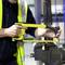 A person in a yellow hi-vis vest is using a hacksaw to cut metal. Workshop elements are blurred in the background.