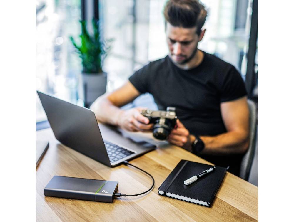 A man sits at a desk with a laptop, holding a camera. Next to him lies a notebook with a pen. A plant is in the background.