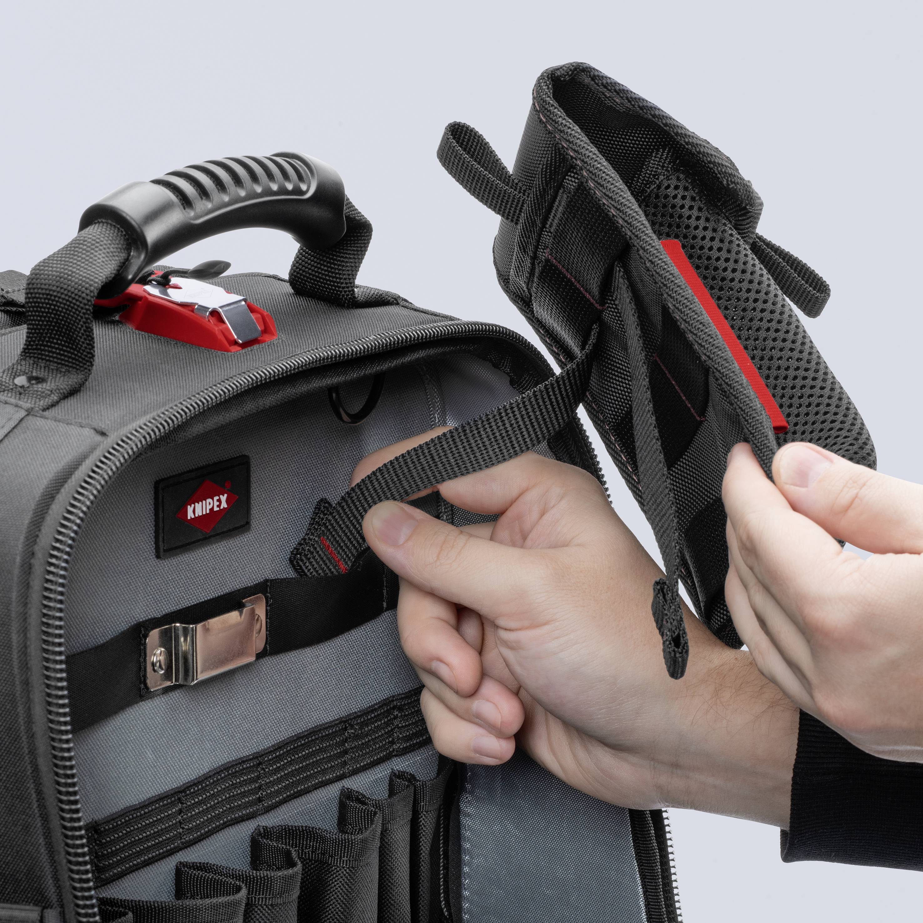 An open tool bag with multiple compartments, with a hand lifting one compartment. The bag is black with red accents.