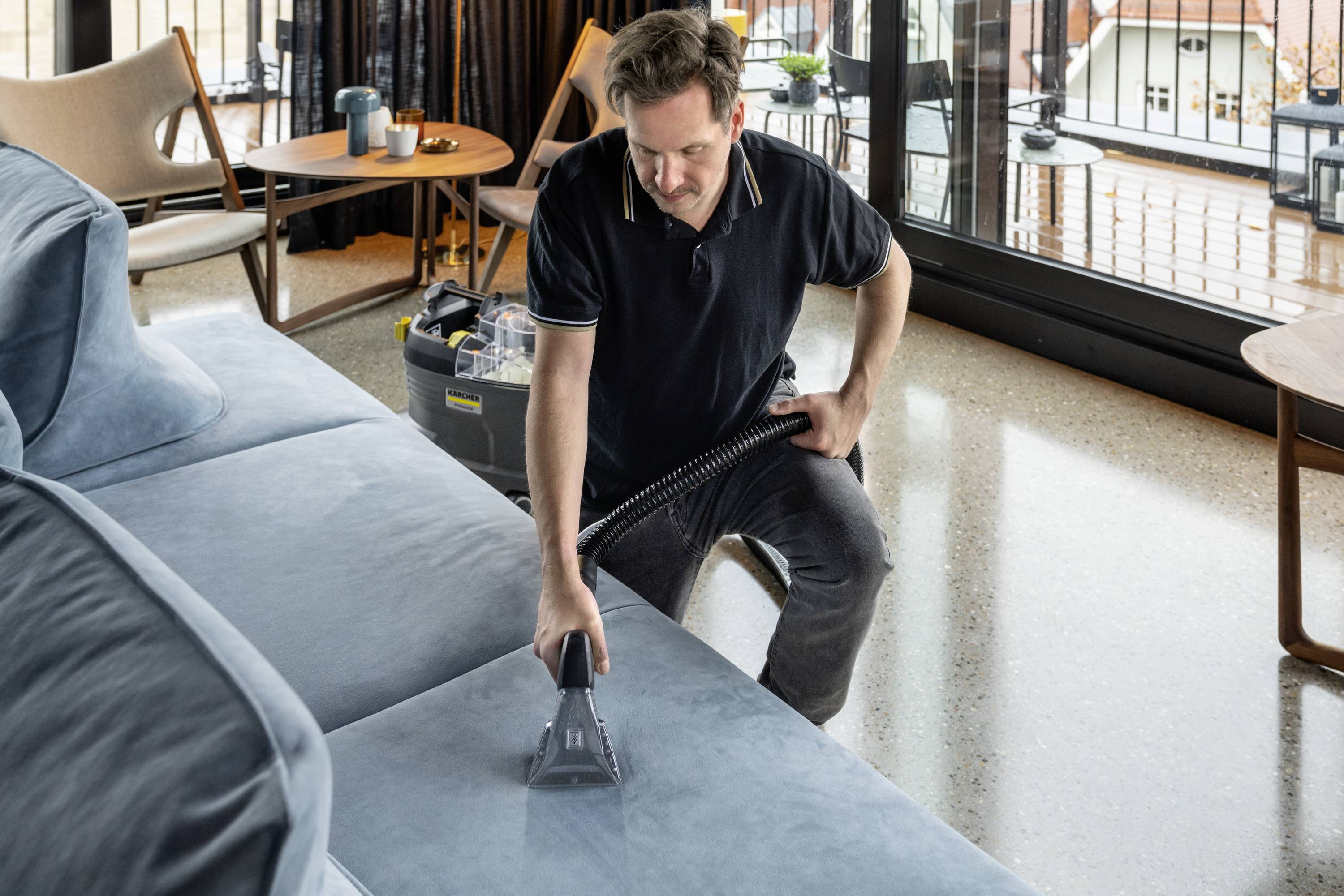 A man is cleaning a blue sofa with an upholstery cleaning device in a living room with large windows.