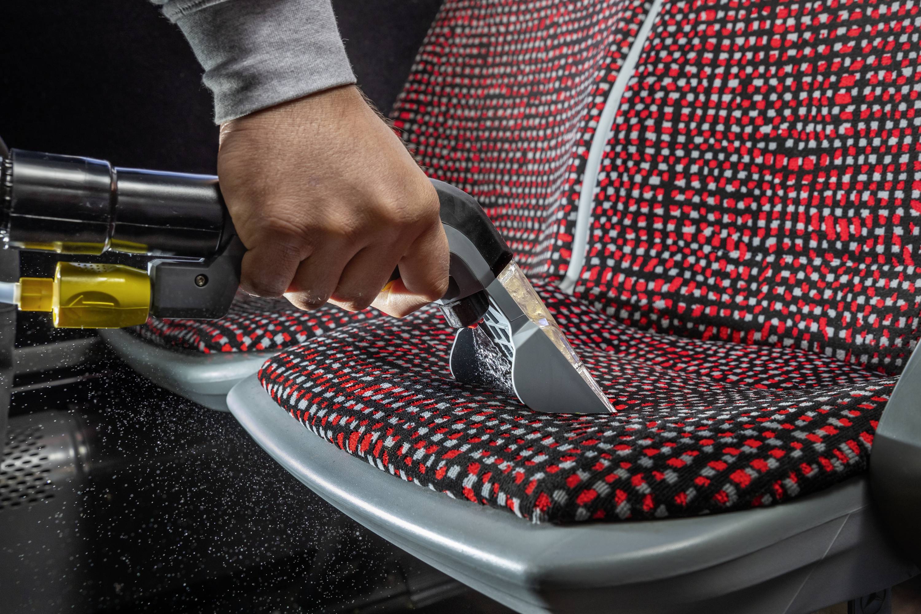 A person is cleaning a red and black patterned seat with a handheld vacuum cleaner. Water droplets are spraying from the nozzle.