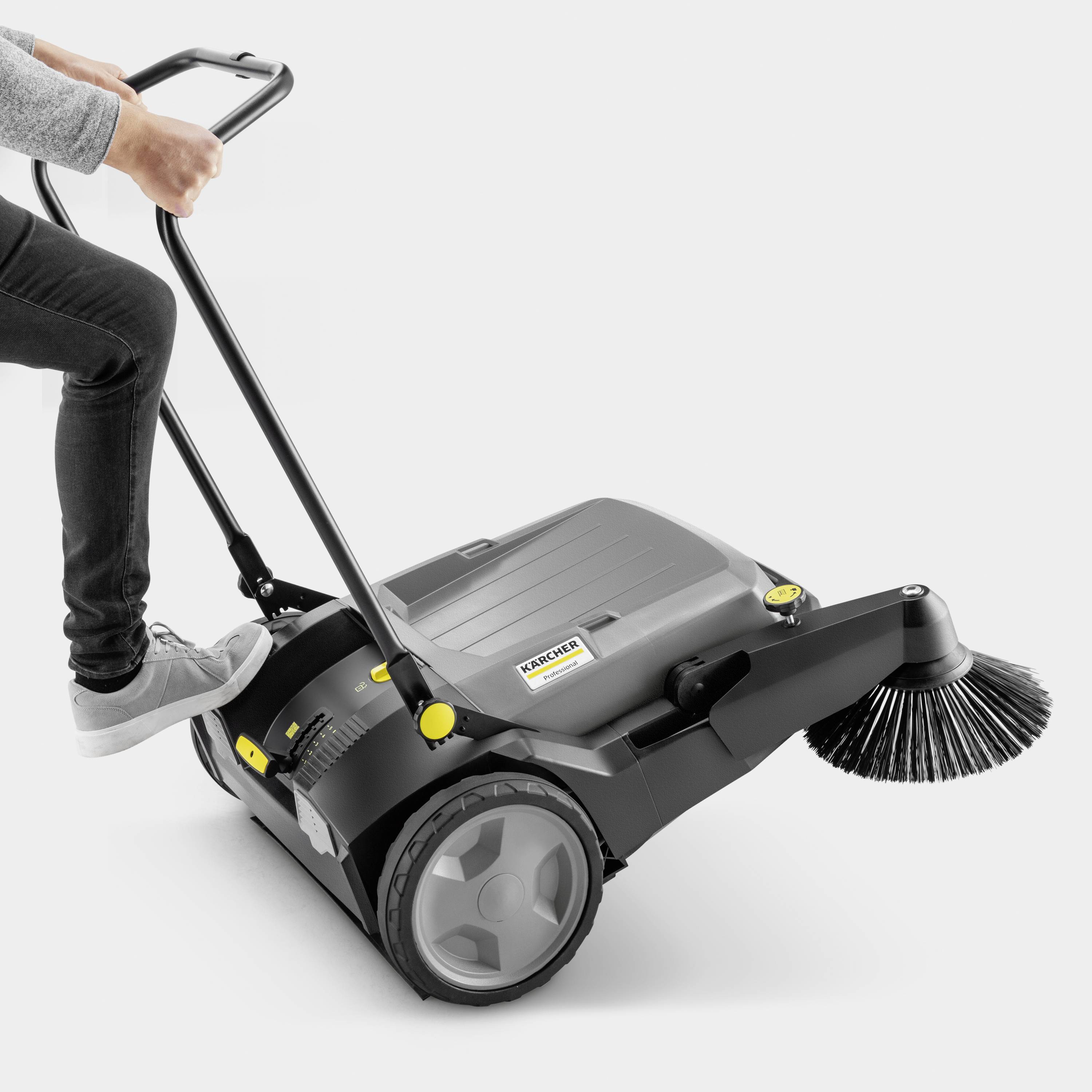 A person is operating a sweeping machine on a flat surface. The machine has two large wheels and a side brush.