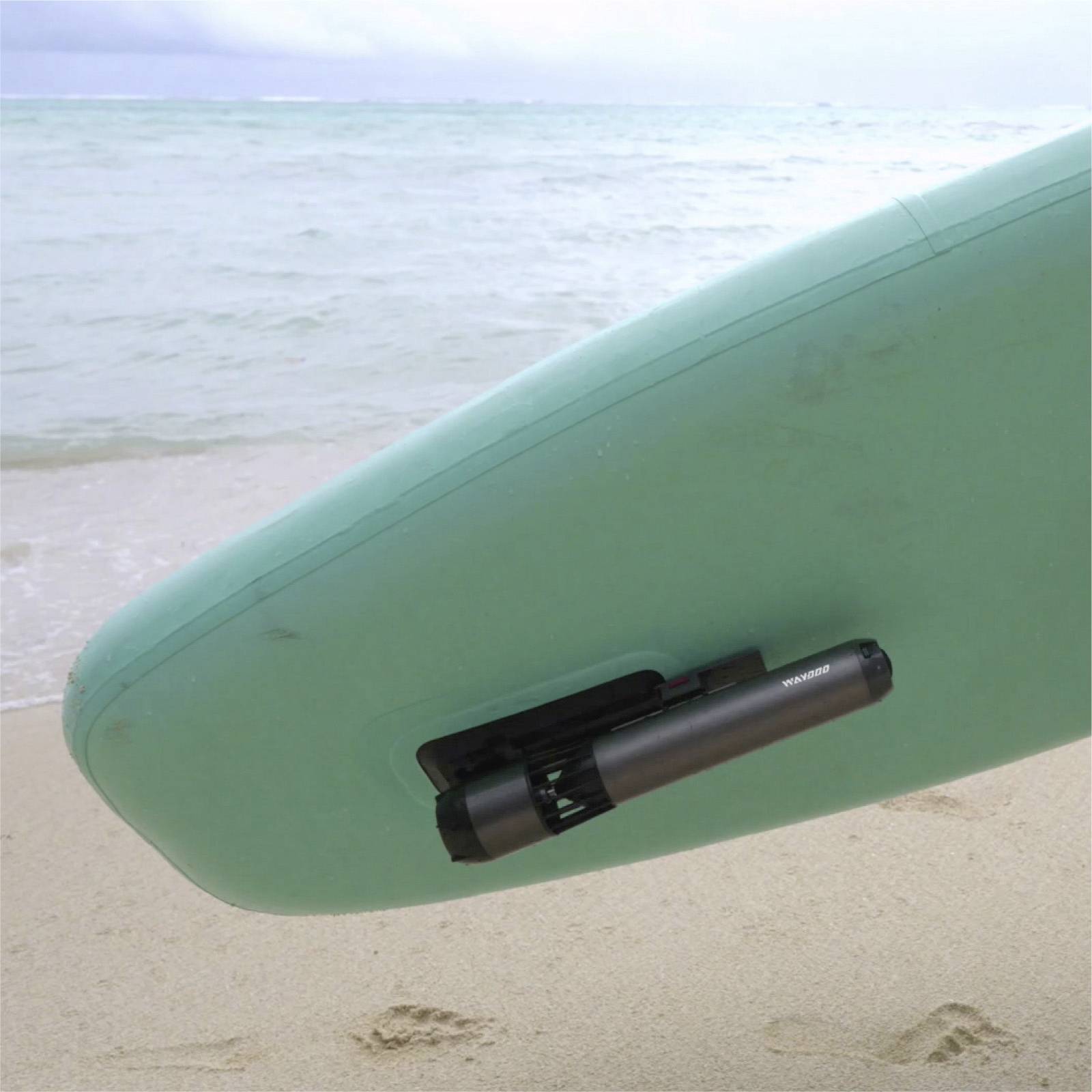 A green surfboard stands upright on the beach with the sea in the background. A black object is attached to the board.