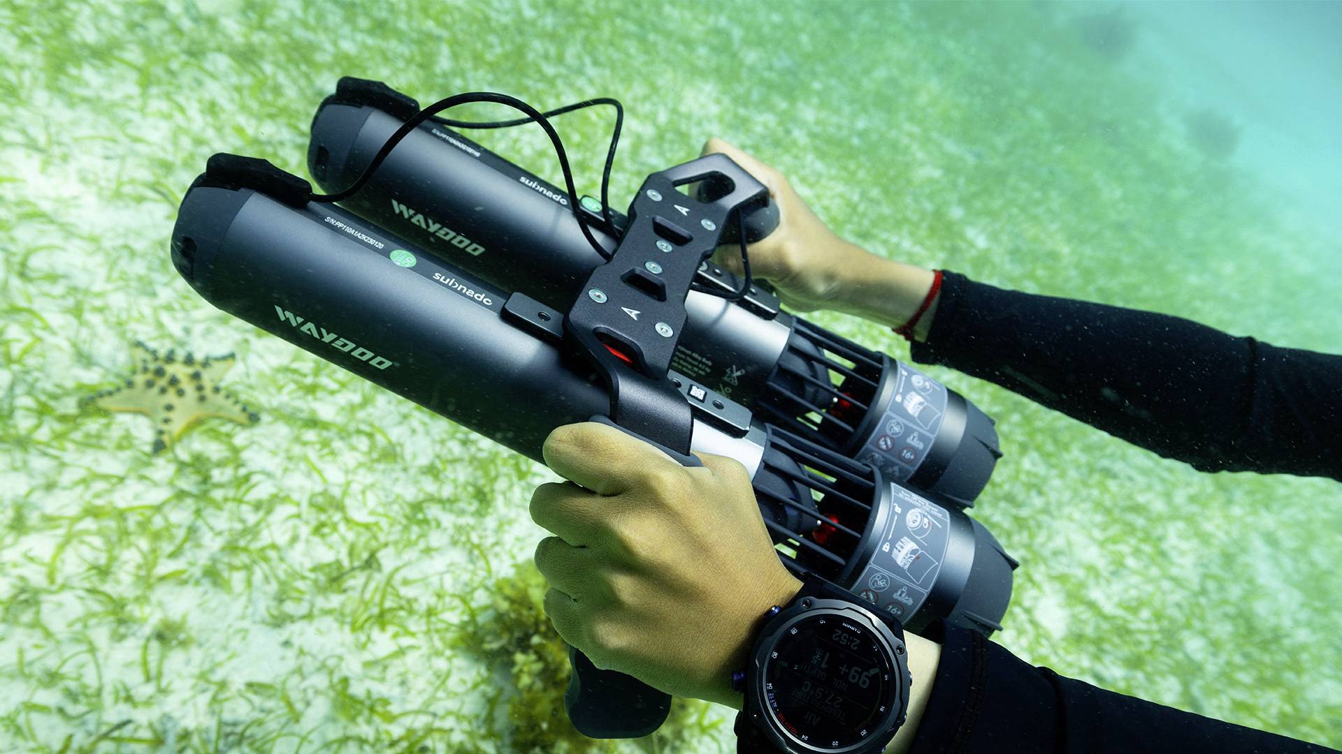Two hands are holding an underwater scooter device in the sea, with a seabed covered in grass and a starfish in the background.