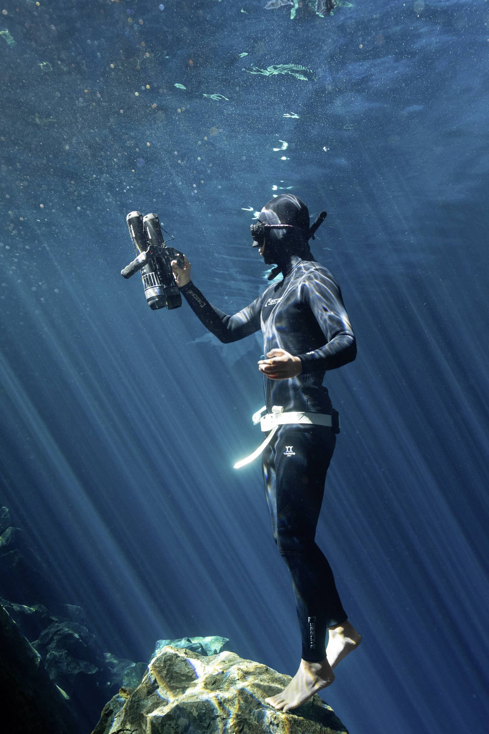 A person in a diving suit stands on a rock underwater, holding a camera, surrounded by rays of light in the clear water.