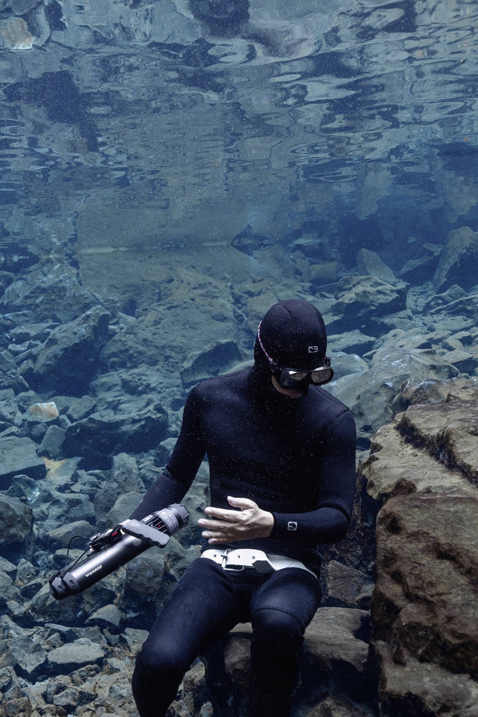 A diver in a black wetsuit and mask is holding a camera underwater, surrounded by rocks and clear water.