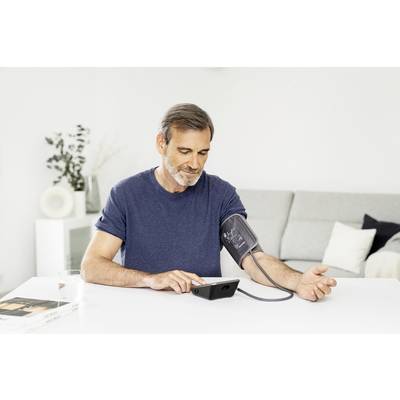 Medieval man measuring his blood pressure with a digital blood pressure monitor, sitting at a table in the living area.