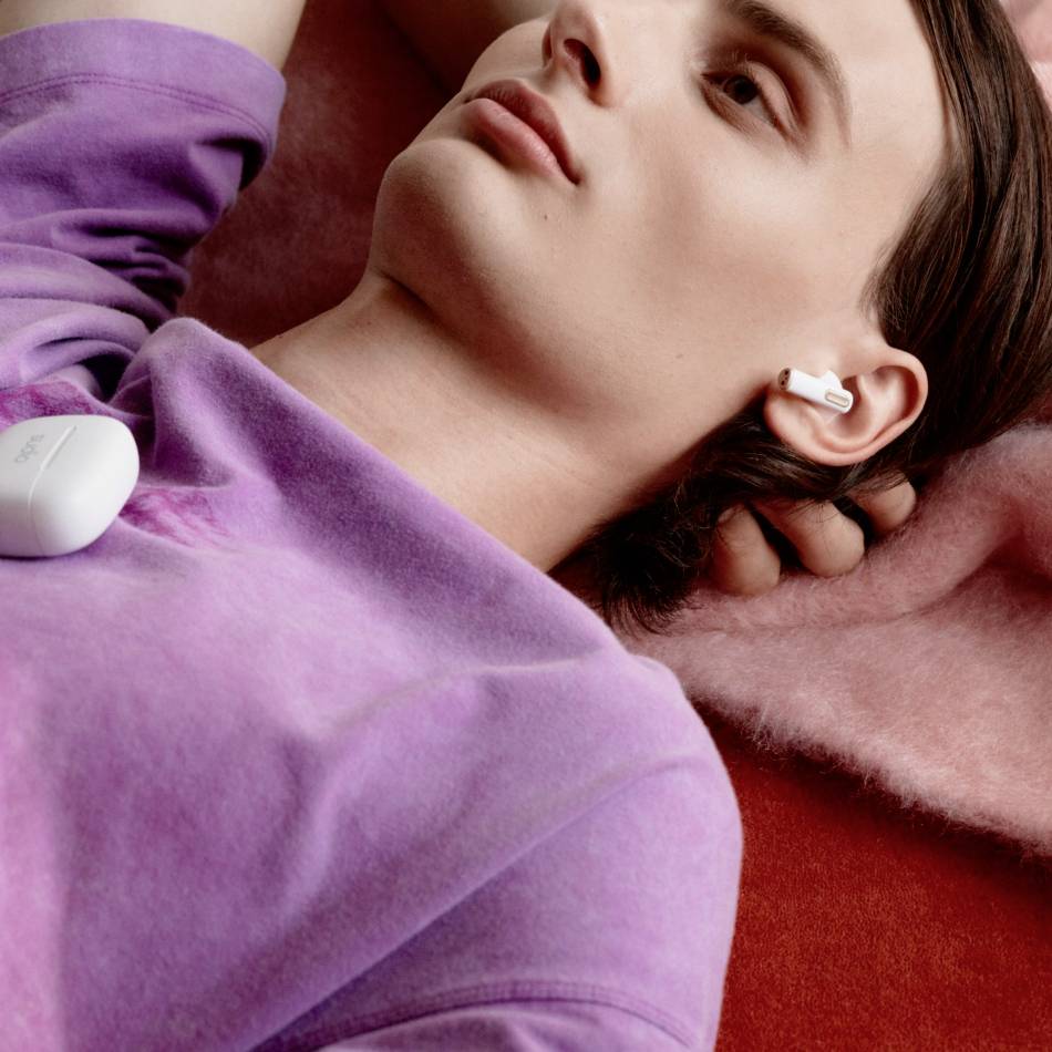 A person is relaxing on a pink blanket, wearing a lilac top and white wireless earphones, with an earphone case beside them.