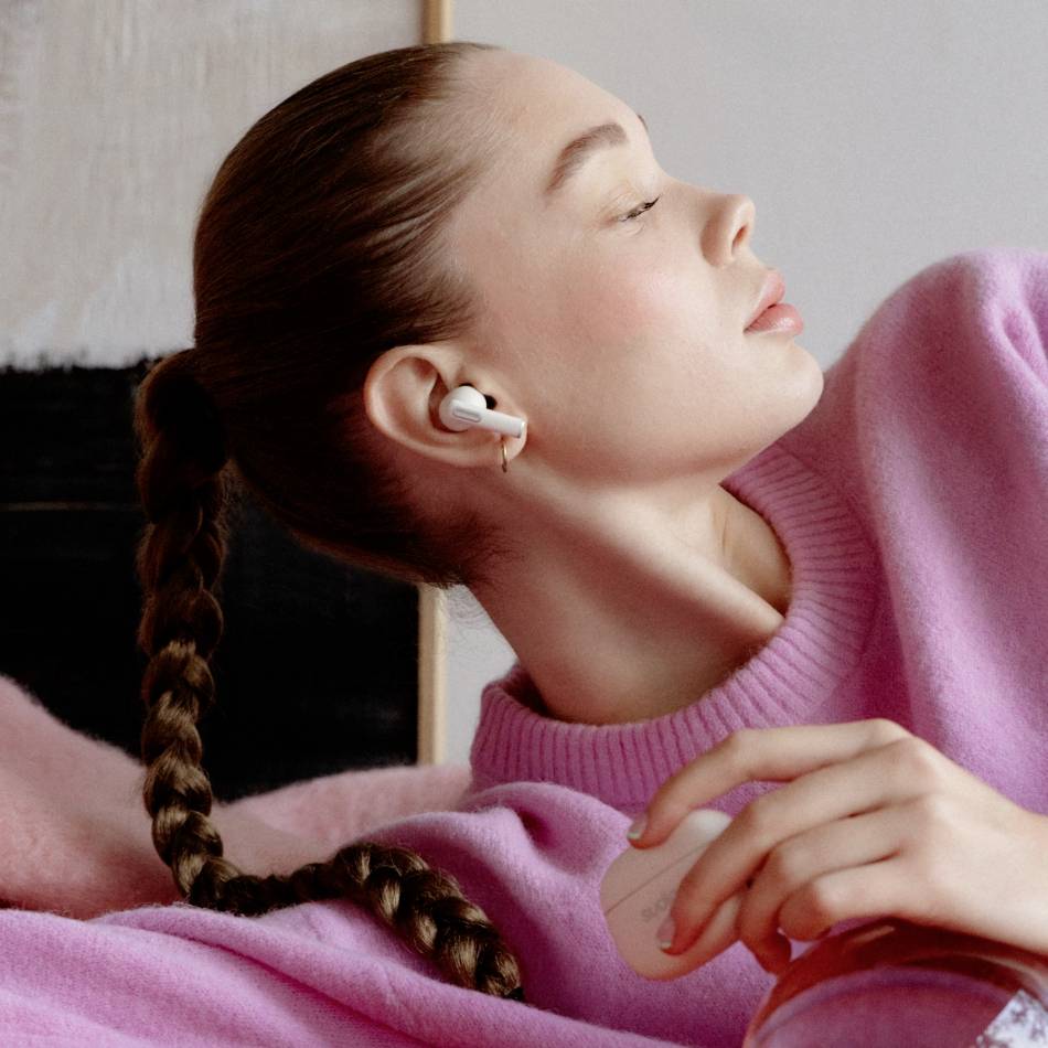 A woman wearing headphones and a pink jumper leans back and enjoys music, in a relaxed pose.