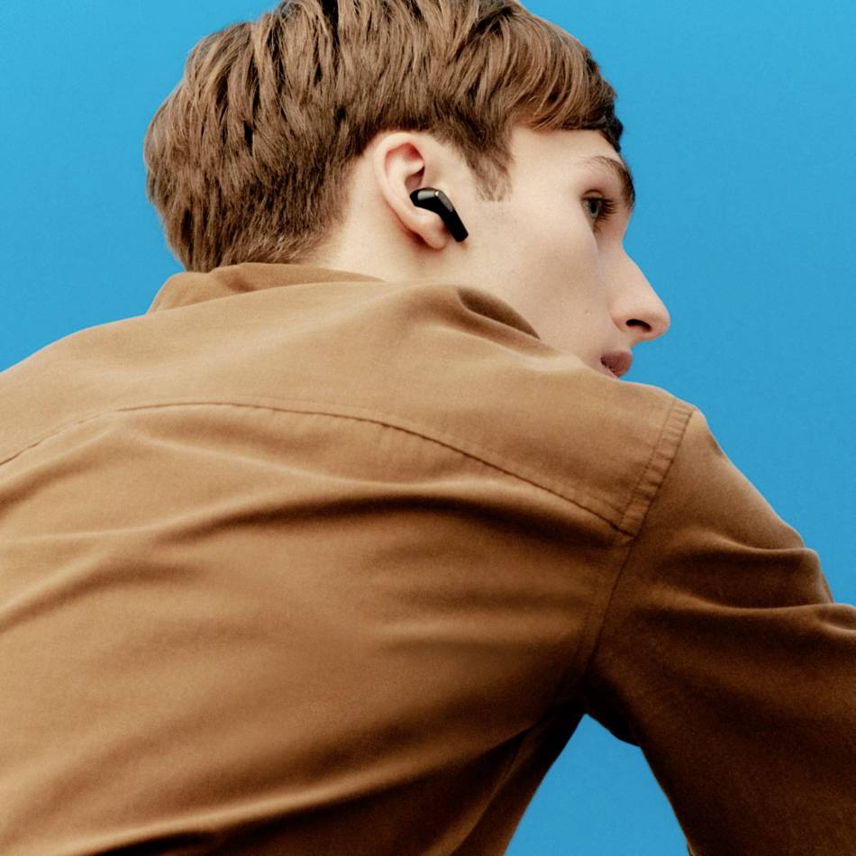A person wearing a brown shirt is wearing black wireless earphones and looking sideways in one direction; blue background.