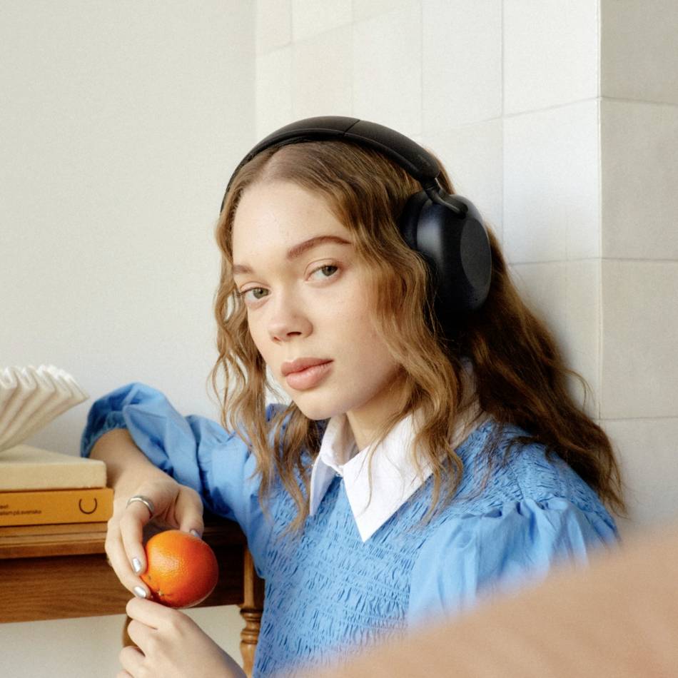 A woman is wearing headphones and holding an orange while sitting at a table and looking into the camera.