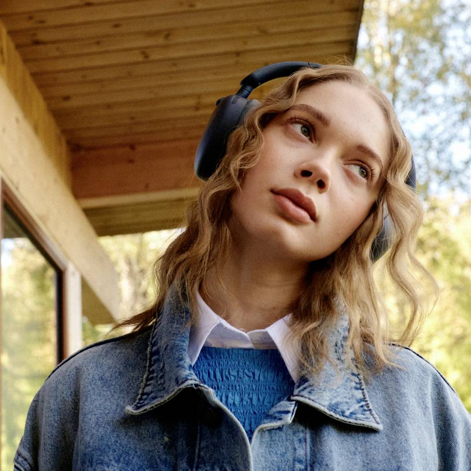 A person with long hair is wearing headphones and a denim jacket, looking relaxed and gazing upwards, standing in front of a wooden-clad building.