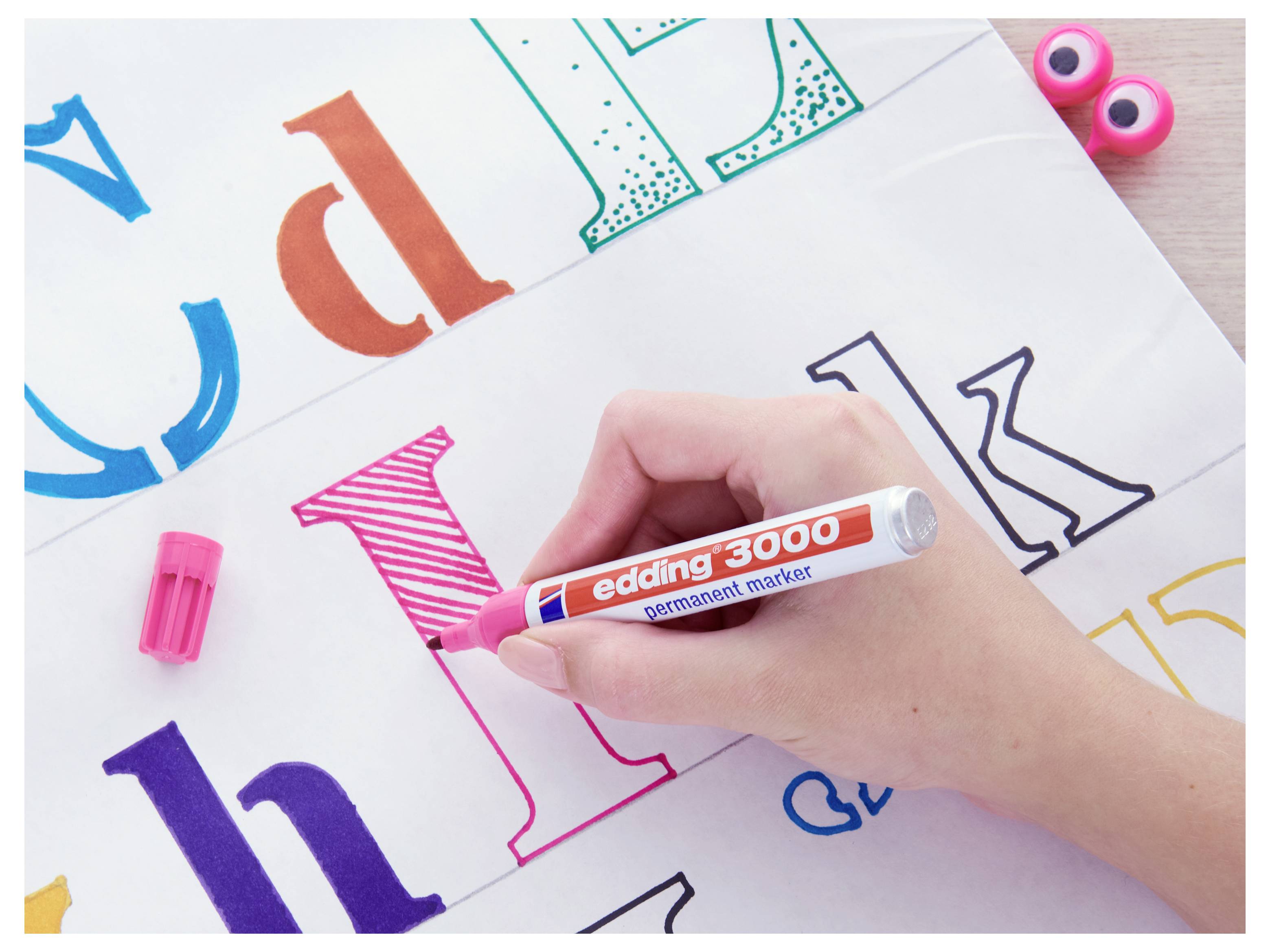 Hand using a pink marker to color a large outlined letter I among colorful alphabet letters on paper.<br>