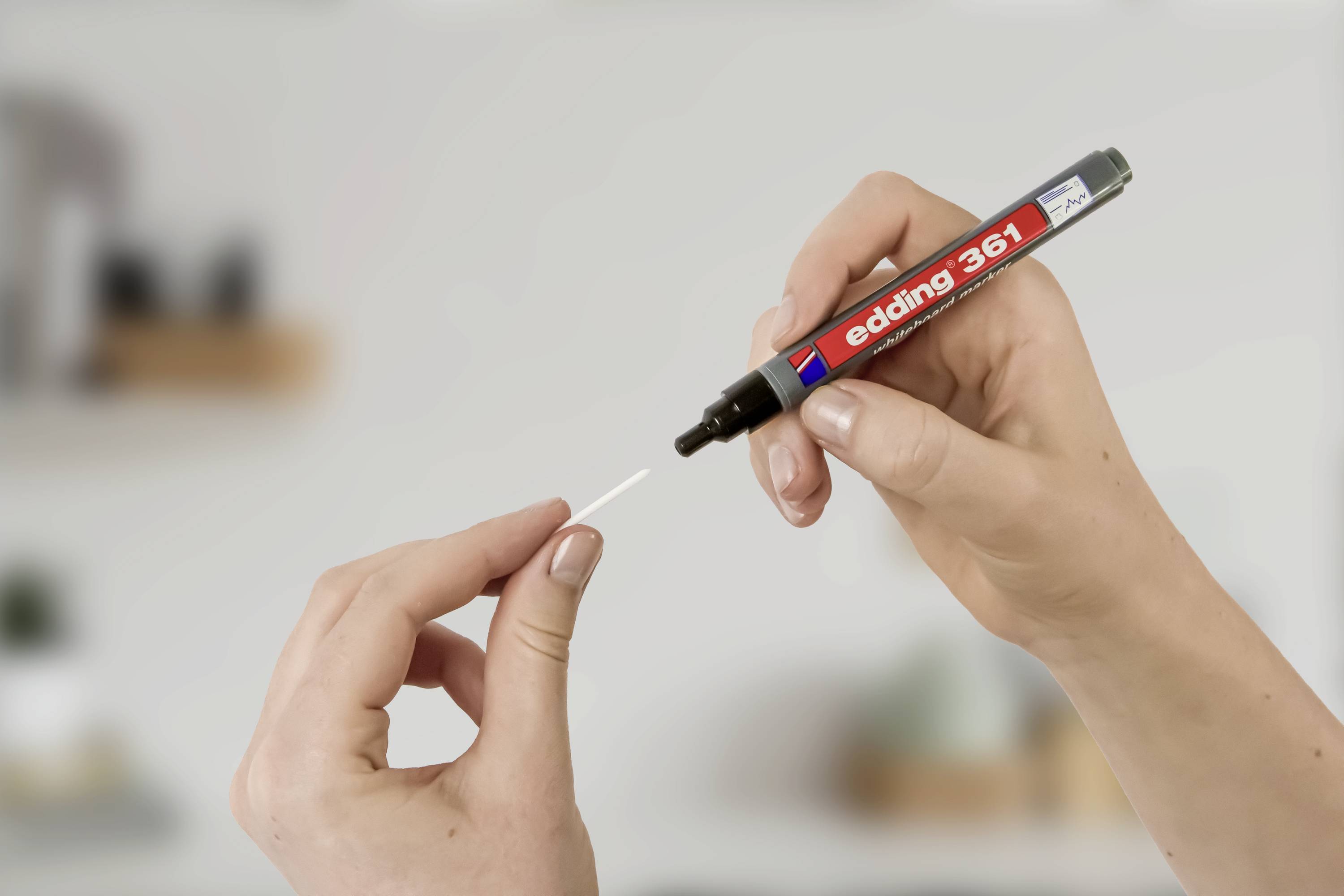 Two hands are holding a red marker and a cotton bud. The marker is hovering over the cotton bud, ready to draw.