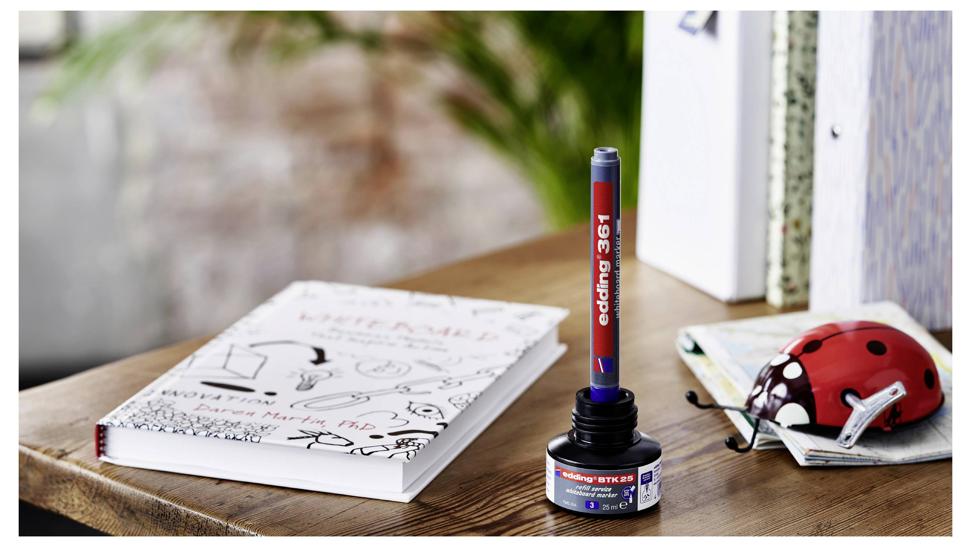 Red and blue 'e' stamp pen standing on a wooden table beside a notebook and a red ladybug-shaped toy car.