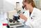 A woman in a white lab coat is using a microscope in a laboratory. Glass bottles and laboratory equipment can be seen in the background.