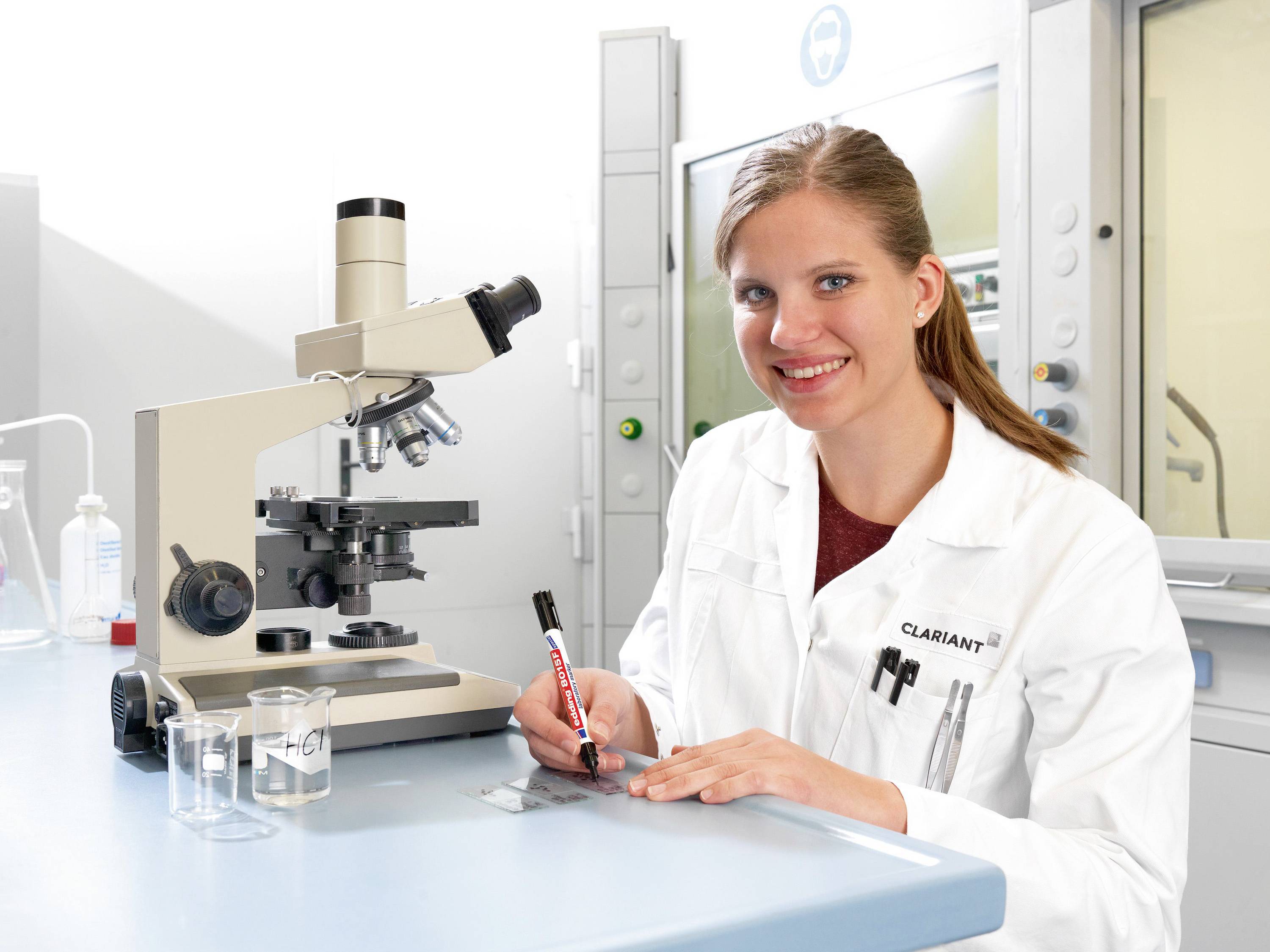 A woman in a white lab coat is working in a laboratory with a microscope and pens. She looks cheerful and is surrounded by laboratory equipment.
