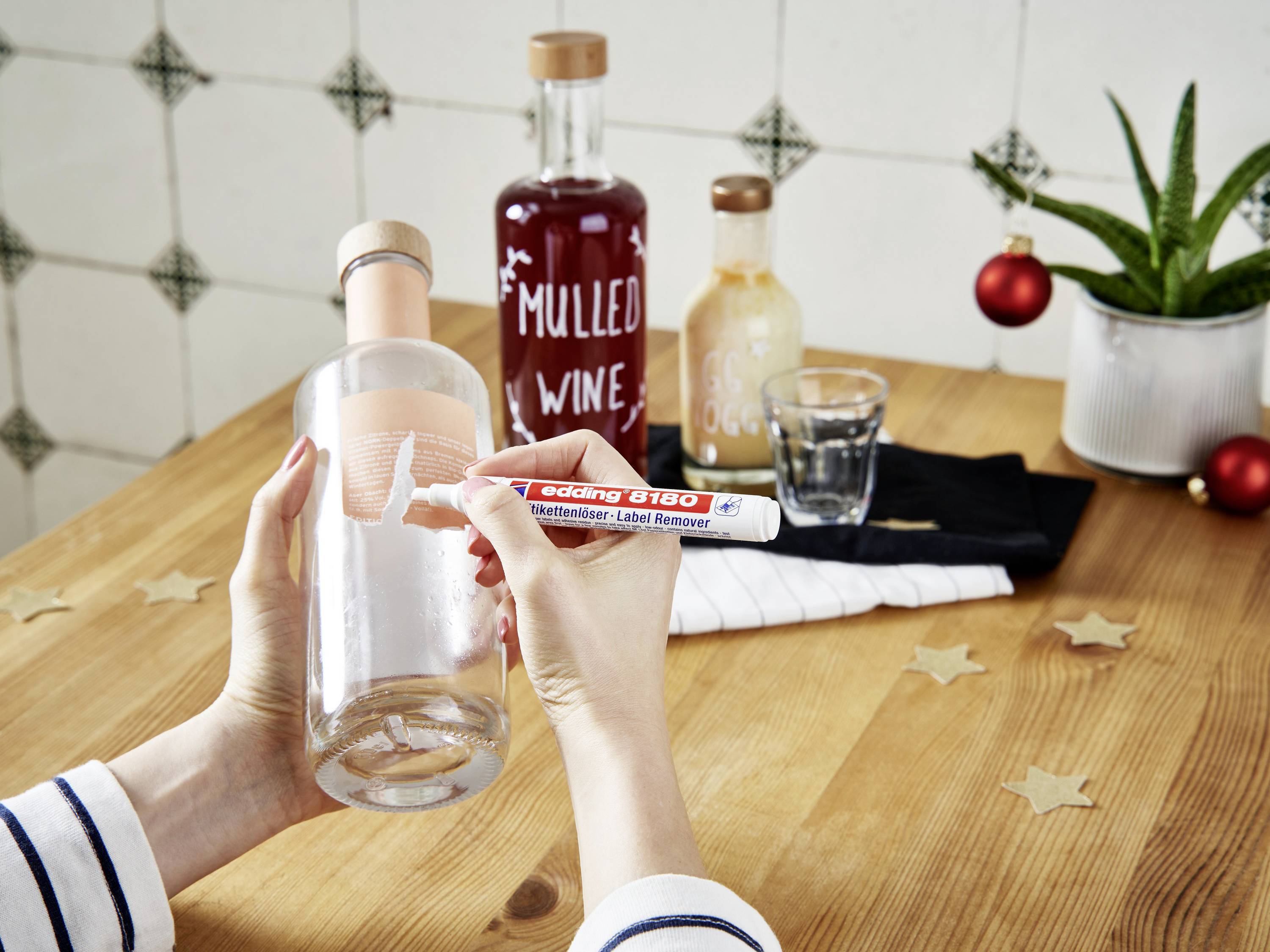 A person is removing a label from a glass bottle using a label remover pen. In the background, additional bottles are placed on a table.
