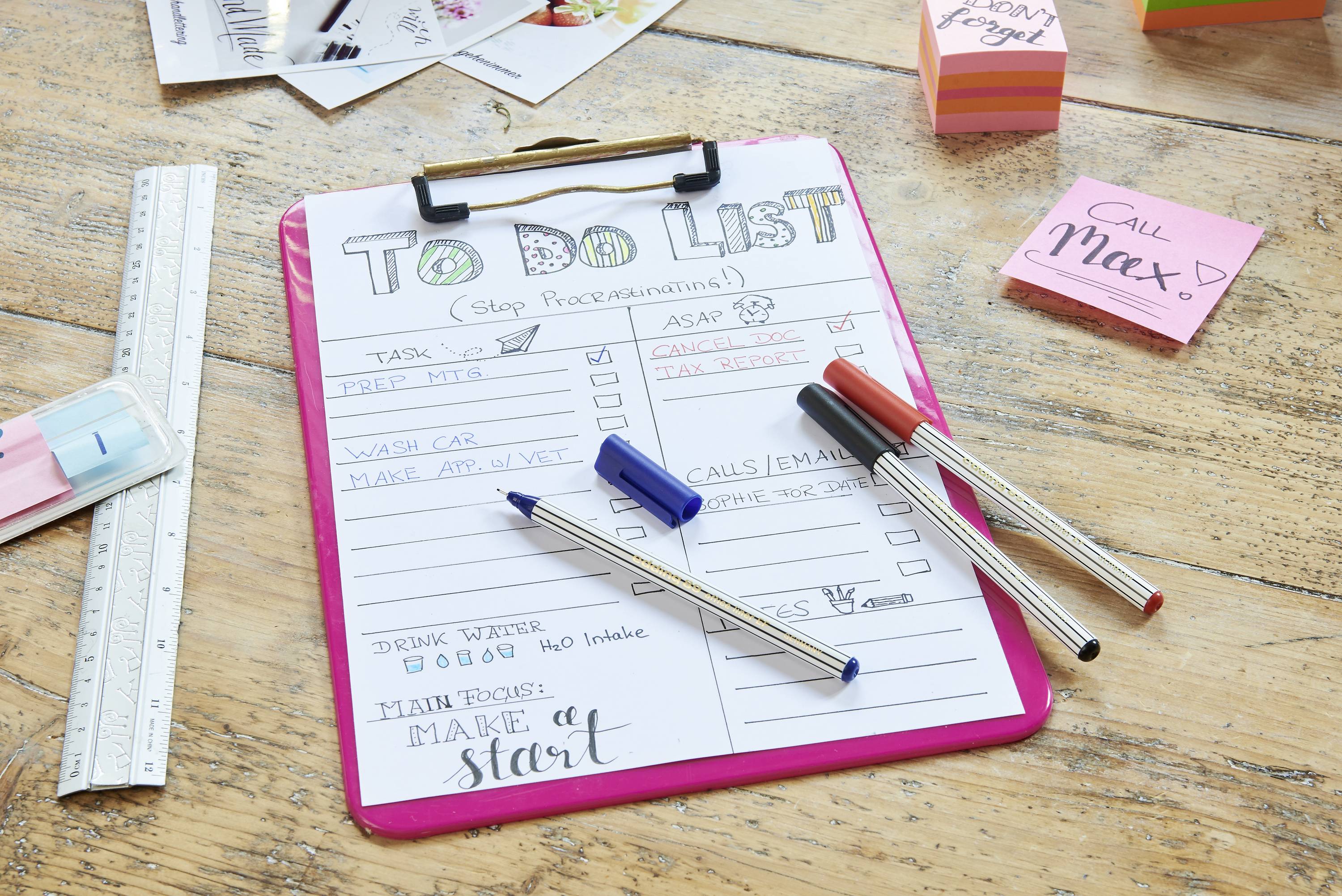 Clipboard on a wooden table with a handwritten to-do list and pens. Notable items: 'Wash car', 'Calls/Emails'.