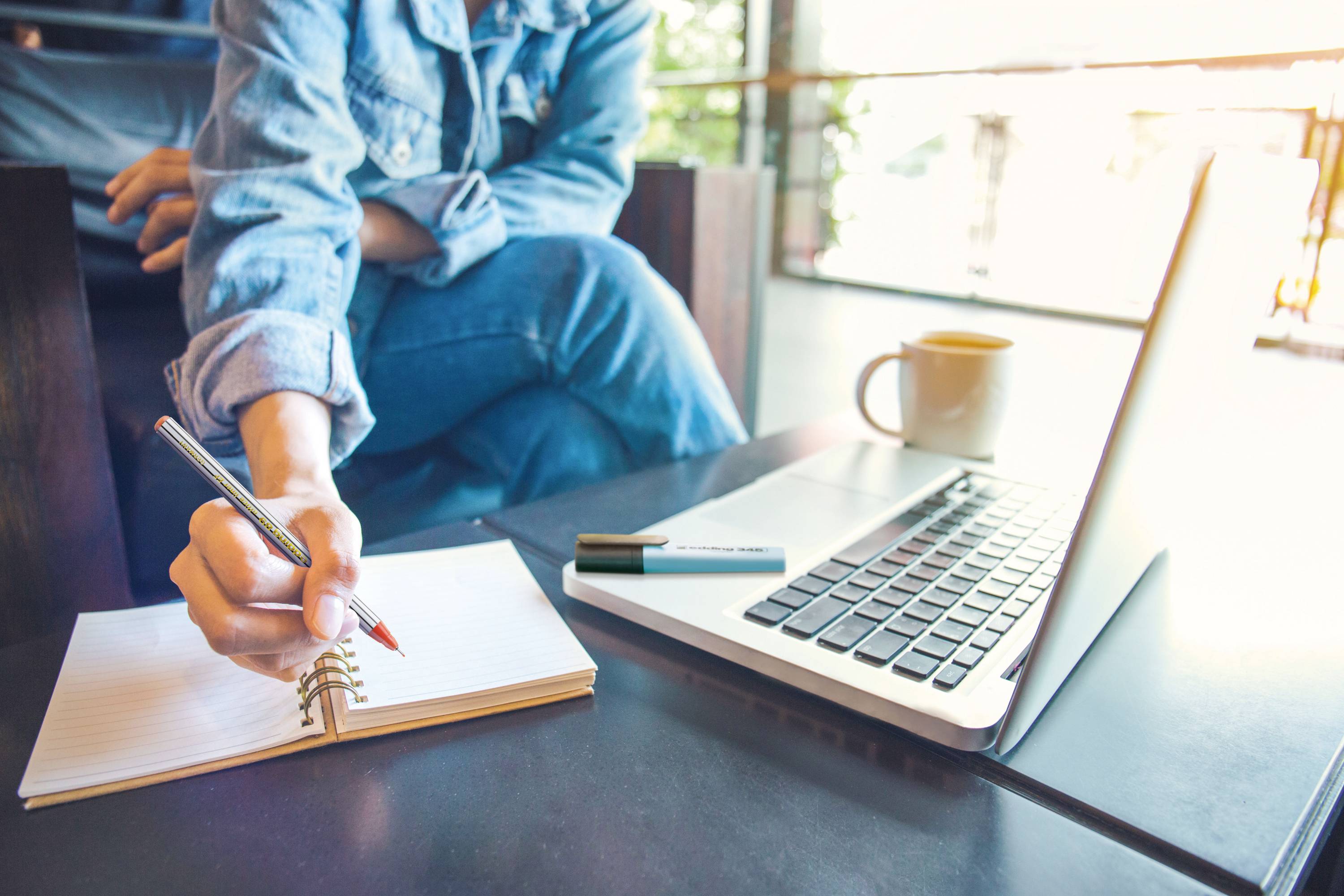 A person is sitting at a table and writing in a notebook, with a laptop and a cup beside them.