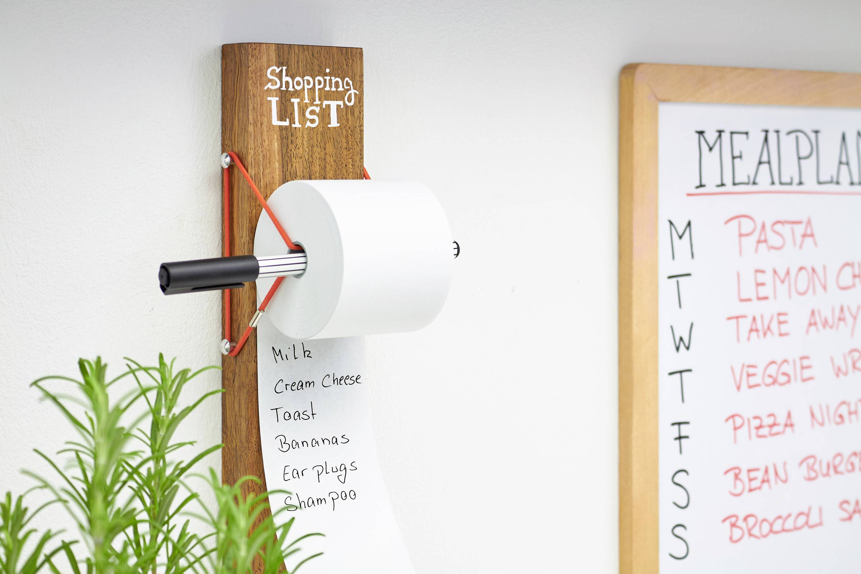 A wooden wall mount with a rolled-up shopping list, noting: milk, cream cheese, bread, bananas, earplugs, shampoo.