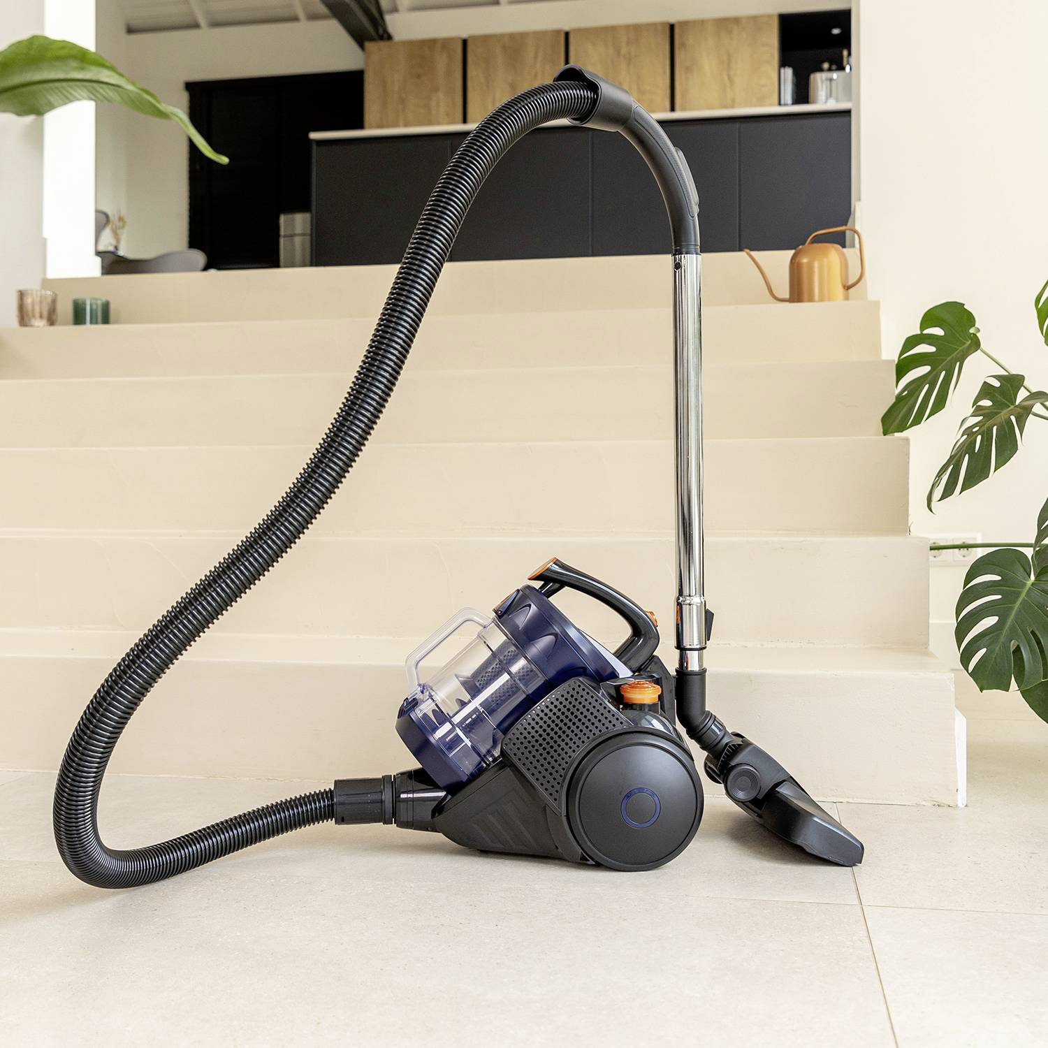 A vacuum cleaner stands on a tiled floor in a modern room. Kitchen cupboards and plants are visible in the background.