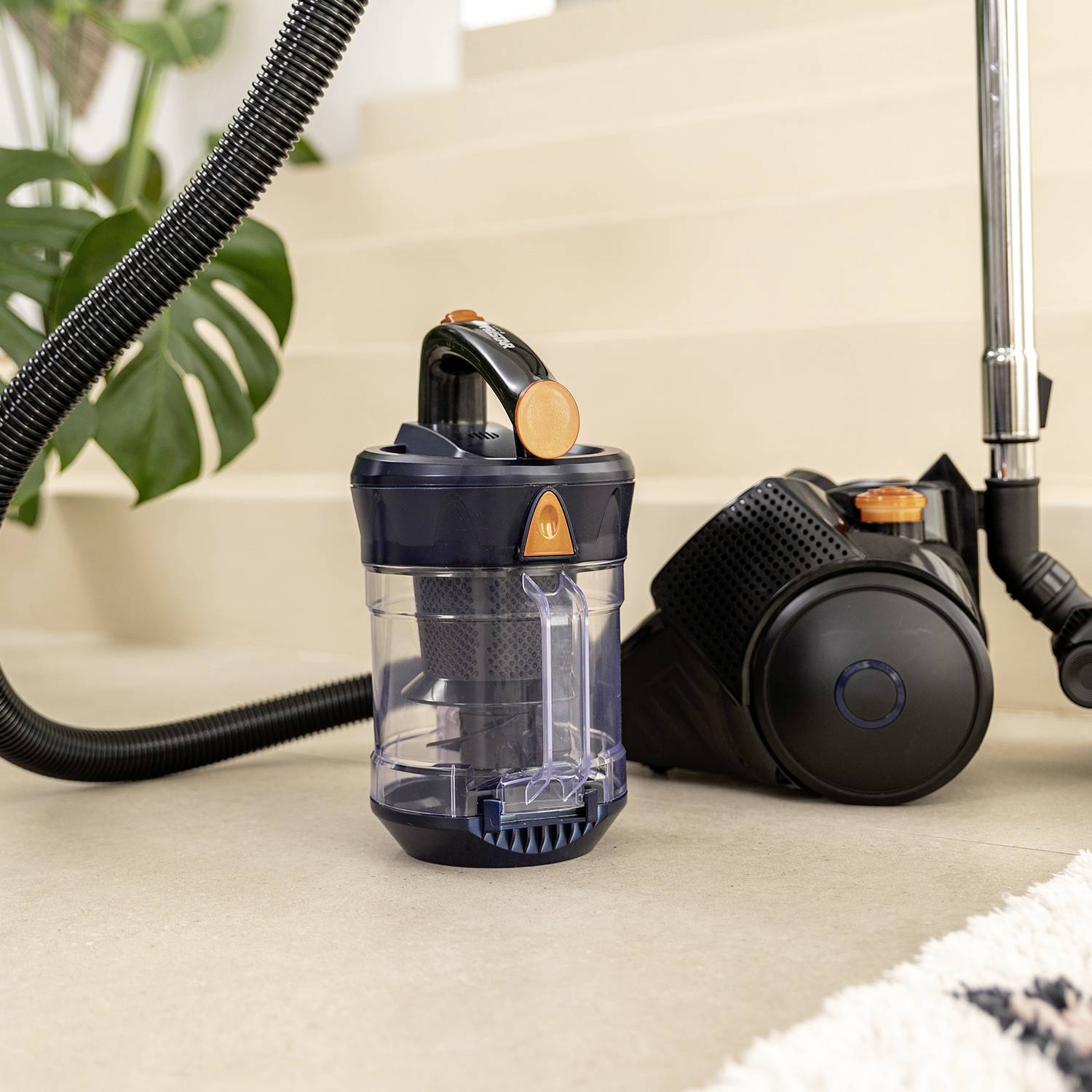A bagless vacuum cleaner is standing on a staircase, with a transparent container in the foreground. A plant is visible in the background.