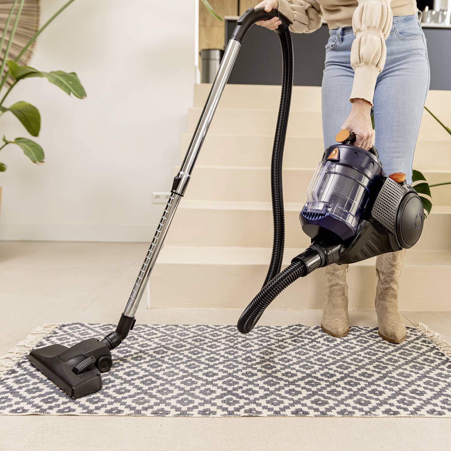 A person is vacuuming a carpet with a black and blue hoover. Only legs and the appliance are visible, in a modern living environment.