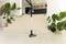 A cordless vacuum cleaner stands on a light floor in front of a staircase in a modern room with large indoor plants.