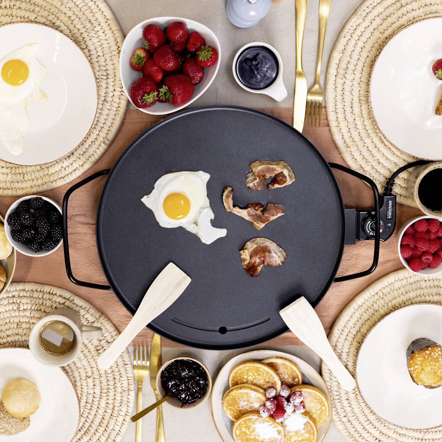 A table with a pancake griddle in the centre, surrounded by plates of fried eggs, bacon, berries, jam, and cutlery.