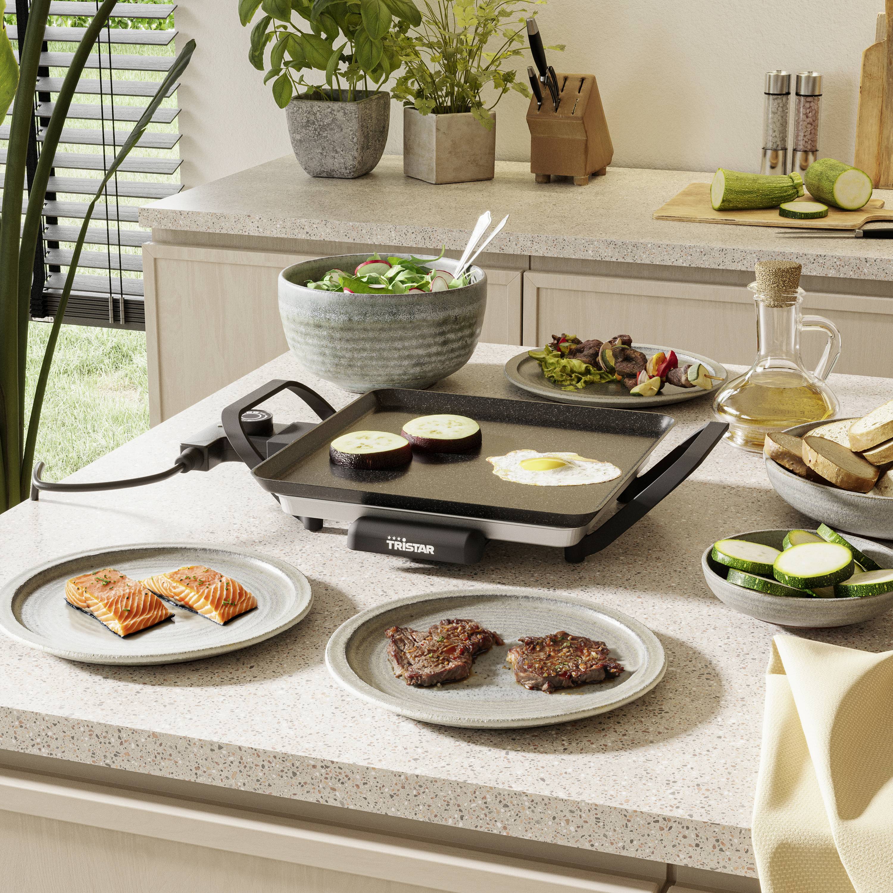 A kitchen with an electric griddle. Meat slices and vegetables are being grilled on it. Plates with salmon and bread slices are lying on the counter.