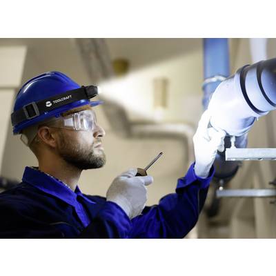 A tradesman wearing safety glasses and a hard hat is inspecting a pipe with a torch in an industrial setting.