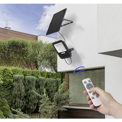 A hand is holding a remote control that activates a solar-powered outdoor light on a house. A garden and blue sky are visible in the background.