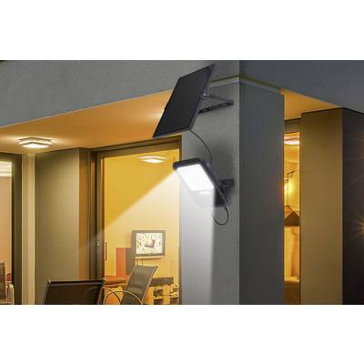 A solar lamp is fixed to a house wall and illuminates the area beneath it. In the background, a lit room is visible.