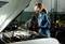 A person in blue workwear is inspecting an open engine compartment, holding a torch and wearing safety glasses and gloves.