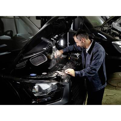 A car mechanic is working under the raised bonnet of a vehicle in a workshop, handling tools.