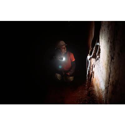 A person wearing a helmet and carrying a torch carefully examines a dark cave or tunnel, illuminating the damp walls with close attention.